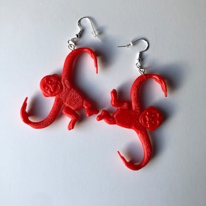 Puede incluir: Un par de pendientes de mono de plástico rojo. Cada pendiente presenta una pequeña figura de mono estilizada con una cola curvada y ganchos para la oreja de color plateado. Los monos son de un color rojo brillante y sólido.