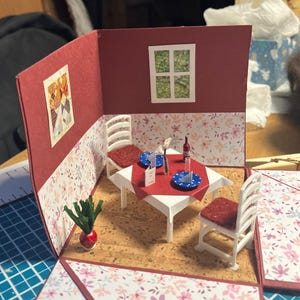 Könnte beinhalten: Eine Miniatur-Puppenhaus-Esszimmerszene mit einem weißen Tisch mit roter Decke, zwei weißen Stühlen mit roten Polstern, einer kleinen Flasche, blauen Tellern und einer Speisekarte. Die Wände haben florale Tapeten, ein Fenster und ein Bild.