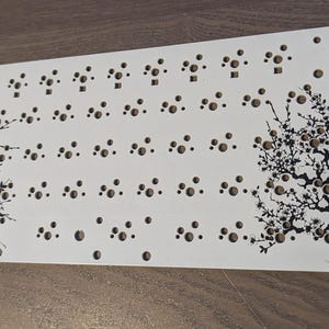 Könnte beinhalten: Weiße Metallplatte mit einem schwarz-weißen Design aus zwei stilisierten Bäumen. Die Platte hat ein Raster von Löchern zum Befestigen von elektronischen Bauteilen. Der Text "Panel Controller Test Board" ist in der oberen rechten Ecke gedruckt.