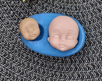 Könnte beinhalten: Eine blaue Silikonform mit zwei Puppengesichtern. Ein Puppengesicht ist hellbraun, das andere blassrosa. Die Form befindet sich auf einer grau-schwarzen gewebten Oberfläche, mit einer gelben Blume im Hintergrund.