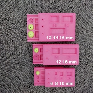 Pode incluir: Três moldes de silicone retangulares cor-de-rosa com várias cavidades quadradas e retangulares. Os moldes apresentam etiquetas numéricas 6, 8, 10, 12, 14 e 16 mm, indicando os tamanhos das cavidades. Várias contas esféricas verde-claras estão colocadas.
