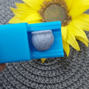 Op de afbeelding: Een kleine, gespikkelde grijze keramische pot staat in een helderblauwe houder. De pot heeft een afgeronde vorm met een kleine opening. Op de achtergrond staat een grote gele zonnebloem, met daaronder een grijs patroon.