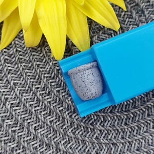 May include: A small grey ceramic pot with a speckled finish is held in a blue plastic dispenser. The pot is approximately 2.5 cm in diameter.