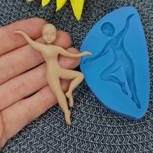 Puede incluir: Una figura de bailarina de color beige en una pose elegante, junto a un molde de silicona azul de la misma bailarina. La figura tiene los brazos extendidos y una pierna extendida, sugiriendo movimiento. El fondo es una tela gris texturizada.