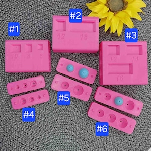 Pode incluir: Nove moldes de silicone rosa para fazer joias. Os moldes são numerados de 1 a 6. O molde nº 1 tem três cavidades retangulares. O molde nº 2 tem duas cavidades retangulares. O molde nº 3 tem três cavidades retangulares. O molde nº 4 tem três cavidades redondas. O molde nº 5 tem duas cavidades redondas. O molde nº 6 tem três cavidades redondas. Os moldes nº 4 e nº 5 têm contas azuis nas cavidades.