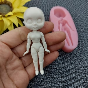 Puede incluir: Molde de plástico blanco para muñeca con una muñeca de plástico blanco en la misma pose. El molde es rosa y tiene una superficie lisa.