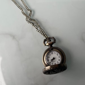 Peut inclure: Pendentif montre de poche de couleur bronze antique avec une chaîne assortie. Le cadran de la montre est blanc avec des chiffres et des aiguilles noirs. La montre est ouverte, révélant l'heure. La chaîne est de conception délicate et à maillons.