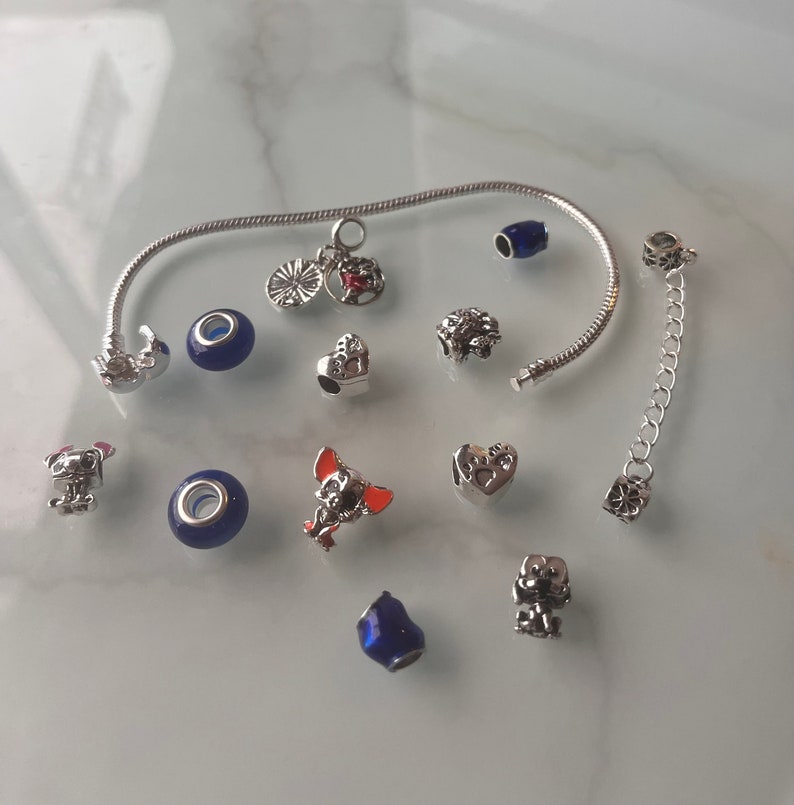 May include: A silver charm bracelet with a variety of charms, including a heart, a dog, a paw print, and a blue bead. The bracelet is on a white surface.