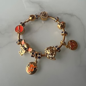 Puede incluir: Pulsera de charms de oro con varios personajes de Disney, incluyendo Simba, Nala y Timón. Los charms son de oro con detalles de esmalte azul y naranja.