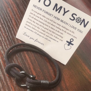Peut inclure: Un bracelet en corde noire avec un fermoir en métal est présenté à côté d'une carte. La carte indique "À MON FILS" et comprend un message d'amour et d'encouragement. Le bracelet est un accessoire sur le thème nautique.