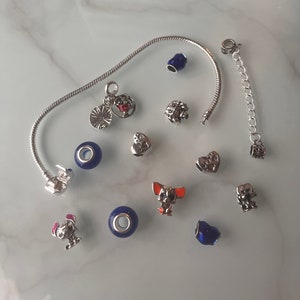 May include: A silver charm bracelet with a variety of charms, including a heart, a dog, a flower, and a blue glass bead. The bracelet is on a white surface.