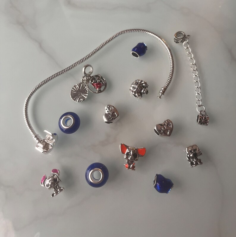 May include: A silver charm bracelet with a variety of charms, including a heart, a dog, a flower, and a blue glass bead. The bracelet is on a white surface.