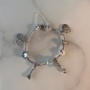 Könnte beinhalten: Ein silbernes Charm-Armband mit verschiedenen Anhängern, darunter Schneeflocken, Herzen und runde Charms. Das Armband mit Kettenverschluss wird auf einer weißen Marmoroberfläche präsentiert. Die Charms sind mit klaren und blauen Akzenten versehen.
