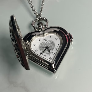 Puede incluir: Un reloj de bolsillo plateado en forma de corazón con una cadena. La esfera del reloj es blanca con números y manecillas negras, y un corazón brillante en el centro. El reloj está abierto, mostrando la hora. Sobre un fondo blanco.