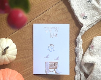 Puede incluir: Una tarjeta de felicitación rectangular blanca con el texto en alemán Willkommen auf der Welt en letra elegante. La ilustración muestra una cuna, un móvil colgante y dos cigüeñas volando en tonos pastel de azul y marrón sobre la tarjeta.