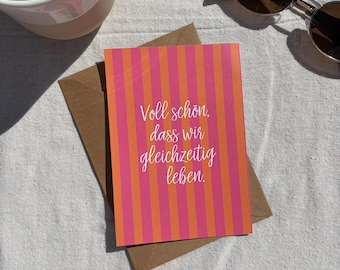 May include: A rectangular greeting card with vertical pink and orange stripes featuring the white cursive text "Voll schön, dass wir gleichzeitig leben." The card is placed on top of a brown paper envelope.