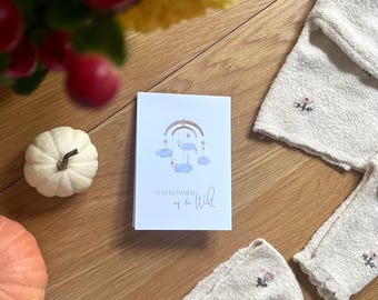 Puede incluir: Una tarjeta de felicitación con una ilustración de móvil para bebé y el texto "Willkommen auf der Wild". La tarjeta está sobre una superficie de madera, con una calabaza blanca, bayas rojas y una toalla crema con bordado floral.