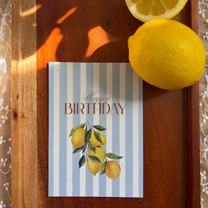 May include: A rectangular birthday card featuring a light blue and white vertical striped background. The text "Happy BIRTHDAY" is printed in brown. Below the text is a watercolor illustration of a cluster of yellow lemons with green leaves.