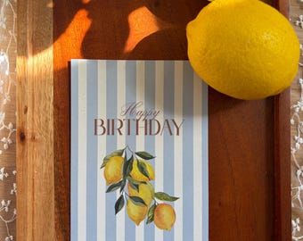 Puede incluir: Una tarjeta de cumpleaños rectangular con un fondo de rayas verticales azul claro y blanco. El texto "Happy BIRTHDAY" está impreso en marrón. Debajo del texto hay una ilustración en acuarela de un racimo de limones amarillos con hojas verdes.