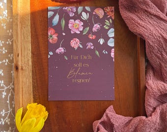 May include: A greeting card with a floral design, featuring pink, purple, and white flowers, and the text "Für Dich soll es Blumen regnen!" on a dark purple background. The card is on a wooden surface with a yellow flower and pink fabric.
