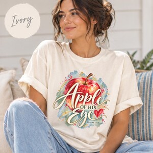 May include: An ivory t-shirt featuring a graphic of a red apple with a watercolor blue and green background. The text reads Apple of His Eye with a small red heart and the scripture reference Psalm 17:8 printed in a script and serif font.