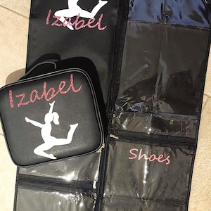 May include: Black garment bag and matching cosmetic case with the name "Izabel" in pink glitter script and a white dancer silhouette. The garment bag has clear pockets labeled "Uniform" and "Shoes".
