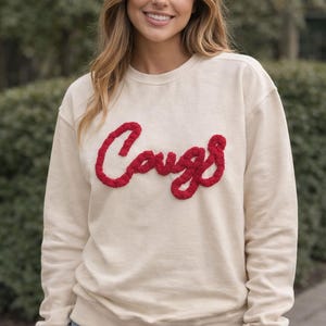 May include: Cream-colored sweatshirt with the word "Cougs" in red, textured lettering. The sweatshirt has a crew neck and long sleeves. The overall style is casual and comfortable.