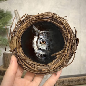 May include: A detailed painting of an owl's face, with striking yellow eyes and white and gray feathers, set within a woven nest. The artwork is a miniature, showcasing fine brushstrokes and a naturalistic style.