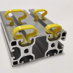 May include: A silver aluminium extrusion profile with four yellow plastic clips attached. The clips are designed to hold cables or wires in place.