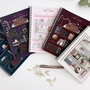 May include: A collection of spiral-bound notebooks with illustrated covers. The notebooks feature bookshelf designs with various decorative elements, including books, plants, and cats. A pen and paperclips are also visible.