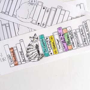 May include: White bookmark featuring a hand-drawn illustration of a cat sitting in front of a bookshelf. The bookshelf includes titles like "Dune" and "The Names." The words "My TBR" are printed on the bookmark.