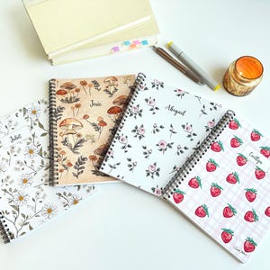 May include: Four spiral-bound notebooks with floral and fruit designs. One notebook features daisies, another has mushrooms, a third has roses, and the last has strawberries on a pink plaid background. Each notebook has a name label.