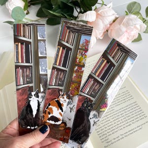 May include: Three clear acrylic bookmarks featuring illustrations of cats in a cozy setting. Each bookmark depicts a different cat breed, with a bookshelf and window in the background. The bookmarks are held near an open book.