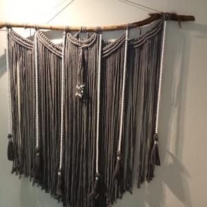 May include: A grey macrame wall hanging with white beads and tassels. The hanging is made of yarn and is attached to a natural branch.