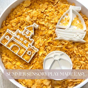Pode incluir: Brinquedos de plástico branco em forma de castelo, barco à vela e bola de praia numa tigela de flocos de milho amarelos. O texto "SUMMER SENSORY PLAY MADE EASY" está na parte inferior da imagem.