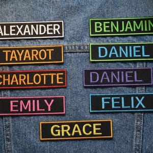 Puede incluir: Parches bordados con nombres en varios colores, incluyendo negro, verde, morado y rosa, se muestran en una chaqueta vaquera. Los nombres incluyen Alexander, Benjamin, Daniel, Emily y Grace.