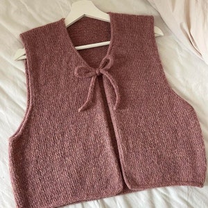 May include: A dusty pink knitted waistcoat with a tie fastening at the neckline.