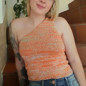 Könnte beinhalten: Nahaufnahme einer Frau, die ein einseitig orange-weiß gestreiftes Stricktop trägt. Das Top ist kurz und hat eine lockere Passform. Die Frau trägt eine blaue Jeans und hat blonde Haare.