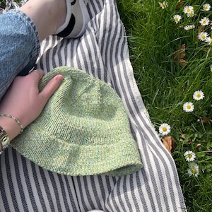 May include: A light green knit bucket hat with a speckled pattern is held on a striped blanket. The blanket is on a bed of green grass with white daisies. A pair of white sneakers and blue jeans are also visible.