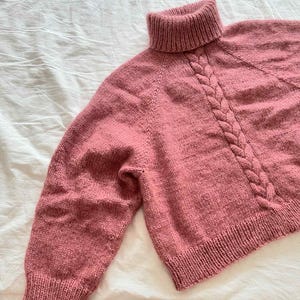May include: A dusty rose-colored knitted sweater with a turtleneck and a cable knit design on the front. The sweater has long sleeves and a ribbed hem and cuffs. The garment is made of a soft, textured yarn.
