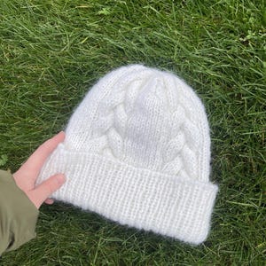 May include: White knitted beanie with a cable knit pattern.