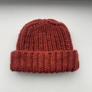 Puede incluir: Un gorro tejido de color óxido. El gorro está hecho de hilo grueso y presenta un borde acanalado. La parte superior del gorro tiene un patrón de tejido apretado. El gorro es un accesorio cálido para el clima frío.