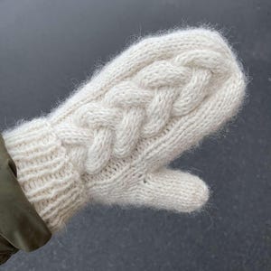 May include: A white knitted mitten with a cable knit pattern. The mitten is made of a soft, fluffy yarn.