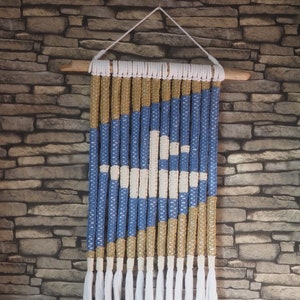 Könnte beinhalten: Ein blauer, weißer und beiger Makramee-Wandbehang mit weißen Quasten. Der Behang besteht aus geknüpften Schnüren und ist an einer Holzleiste befestigt.