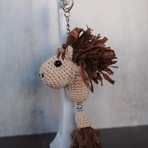 May include: A handmade keychain featuring a crocheted horse head with a brown paper mane and a tassel. The horse is beige with black eyes and a silver-colored keychain ring. The tassel is brown and hangs below the horse's neck.