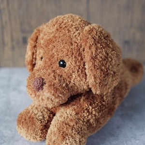 Puede incluir: Un juguete de peluche con forma de perro marrón. El juguete tiene ojos negros y una nariz rosa oscuro. El perro está tumbado, con las patas extendidas. El fondo es una superficie de madera borrosa. El juguete es suave y texturizado.