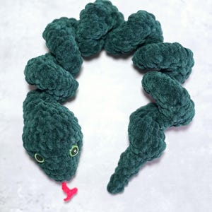 May include: A handmade, plush green snake toy with a red tongue and green eyes. The snake is made of a soft, textured material and is arranged in a curved shape. The toy is designed to resemble a snake.