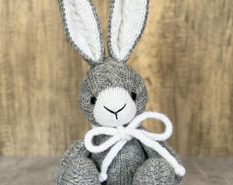 May include: A handmade gray and white knitted bunny rabbit with long ears and a white bow. The bunny has a cable knit pattern on its body and ears. The bunny is sitting upright.