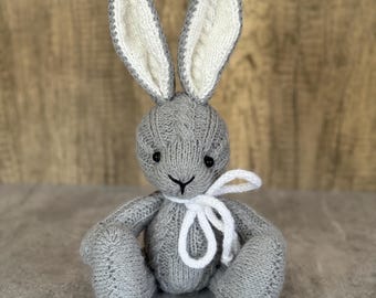 May include: A handmade gray knitted bunny with long ears and a white bow. The bunny's ears have a cable knit design with white inner ears. The bunny has black button eyes and is sitting on a gray surface.