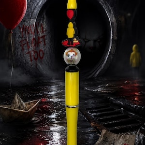 May include: A yellow pen featuring a topper with a figure in a yellow raincoat, a red balloon, and a round bead displaying a clown face. The pen has a silver-colored tip and a yellow barrel, with a design inspired by the horror character Pennywise.